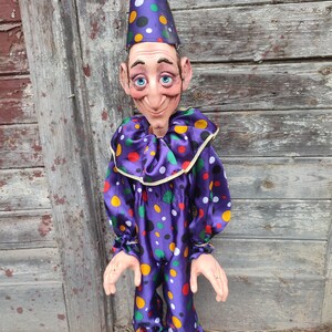 May include: A purple and white polka dot puppet with a purple hat and a painted face. The puppet has long arms and is standing in front of a wooden wall.