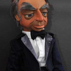 Puede incluir: Un títere con pelo gris, vestido con un esmoquin negro con camisa blanca y pajarita negra. El títere tiene una expresión seria en su rostro.