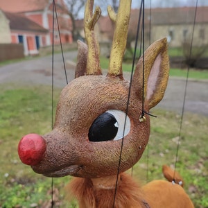 May include: A brown and gold puppet reindeer with a red nose and large black eyes. The reindeer is hanging from strings and has a furry brown body.