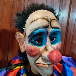 May include: A hand-crafted puppet with a painted face, blue eyes, and a red nose. The puppet is wearing a colorful, multi-striped costume.