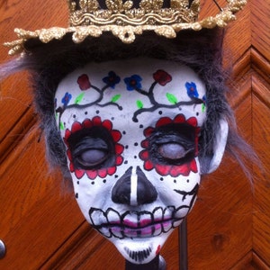 May include: A Day of the Dead puppet with a white face painted with black, red, and blue details. The puppet is wearing a black top hat with gold trim and has gray hair.