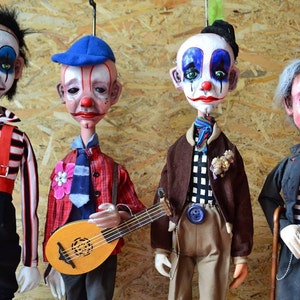 Puede incluir: Cuatro marionetas artesanales vestidas de payasos. Las muñecas llevan ropa colorida y tienen caras pintadas. Una muñeca sostiene una guitarra pequeña.