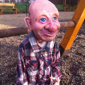 May include: A puppet with a pink face and blue eyes, wearing a plaid shirt. The puppet is hanging from strings and has a cigarette in its hand.