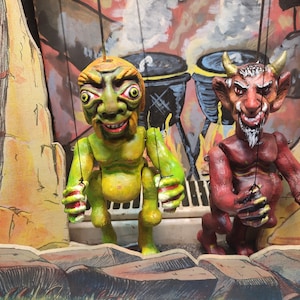 May include: Two puppet figures, one green with large eyes and a toothy grin, the other red with horns and a beard, stand in front of a painted backdrop depicting a fiery scene with cauldrons.