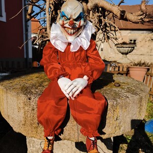 Puede incluir: Una muñeca de payaso aterradora con una expresión amenazante. La muñeca viste un mono rojo, guantes blancos y botas de dos tonos. El payaso tiene un cuello blanco con volantes y una máscara aterradora con detalles azules y rojos.
