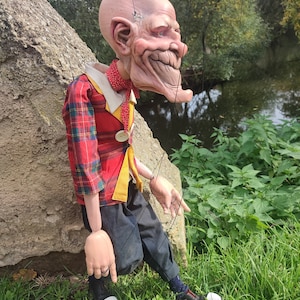 May include: A puppet with a bald head, a red and black plaid shirt, black pants, and white shoes. The puppet has a large, exaggerated smile and is made of a realistic material.