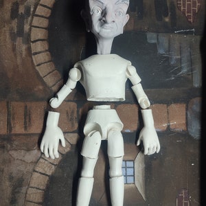 May include: A white, articulated doll with a sculpted head. The doll is disassembled and laid out on a brown surface. The doll's head has a realistic expression with a furrowed brow and a slightly open mouth.