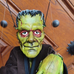 May include: Green Frankenstein puppet with a brown jacket and green hands. The puppet is hanging from a wooden bar with strings.