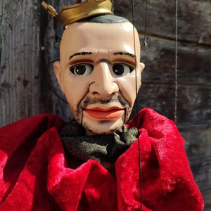 May include: A puppet of a king wearing a gold crown and a red velvet robe. The puppet has a painted face with a mustache and a beard.