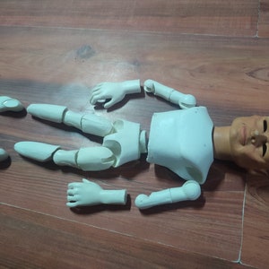 May include: A white plastic doll body with a separate head and limbs. The doll is disassembled and laid out on a wooden surface. The head is painted with a realistic face and has brown eyes.