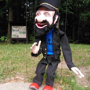 May include: A black and blue puppet with a black hat, a black beard, and red shoes. The puppet is wearing a blue sweater with an anchor design and a black jacket. The puppet is holding its right hand up in a waving motion.
