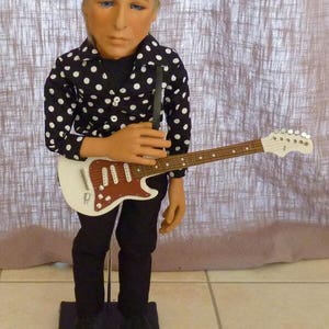 Peut inclure: Une poupée vêtue d'une chemise à pois noirs et blancs, d'un bandeau noir à motif blanc, d'un pantalon noir et de chaussures noires. La poupée tient une guitare électrique blanche avec une touche en bois brun.