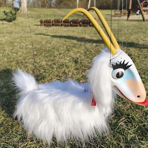 May include: A white puppet goat with a fluffy body, yellow horns, and a painted face with large eyelashes. The goat has a red collar and is suspended by strings, suggesting it is a marionette toy.