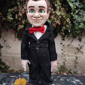 May include: A vintage-style puppet with a painted face, brown hair, and green eyes. The puppet is wearing a black suit with a red bow tie.