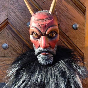 May include: A handcrafted devil mask with a red face, yellow eyes, and light brown horns. The mask has a detailed, gray beard and is surrounded by a black fur collar. The mask is set against a wooden door.