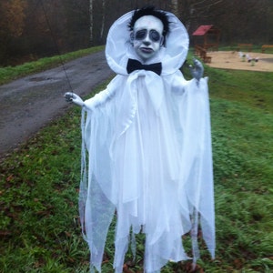 May include: A white ghost costume with a face mask. The ghost has a white collar and a black bow tie. The ghost is standing in a grassy area.