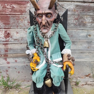May include: A handcrafted puppet with a red and gray face, horns, and a long green coat. The puppet is wearing a chain with bells and is sitting on a black chair.