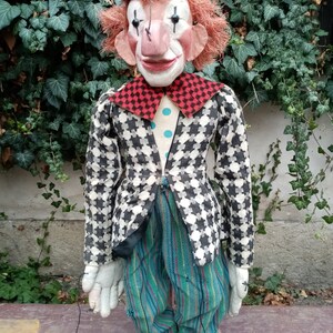 May include: A vintage hand puppet of a clown with a green hat, red hair, and a checkered black and white jacket. The puppet is wearing blue and green striped pants and has a painted face with a red nose and black eyes.