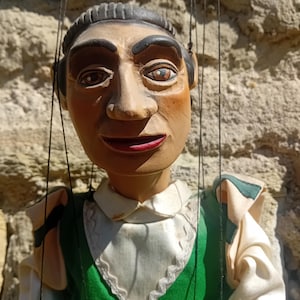 May include: A vintage wooden puppet with a painted face, dark hair, and red lips. The puppet wears a green vest with white trim and a white collar. Black strings are attached to the head and limbs.