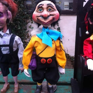 May include: A yellow, blue, and black puppet with a large, smiling face. The puppet has a blue bow tie and is holding a purple rolled-up mat.