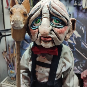May include: A hand-carved wooden puppet with a sad expression, wearing a red bow tie, suspenders, and a button-down shirt. The puppet holds a wooden horse head on a stick. The puppet's face is painted with detailed features.