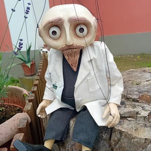 May include: A white-coated puppet with a bald head, large eyes, and a brown beard sits on a tree stump. The puppet is wearing gray pants and black shoes. The puppet is being controlled by strings.