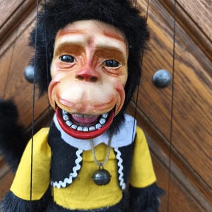May include: A vintage monkey puppet with a smiling face, black fur, and a yellow shirt with a black vest. The puppet has a red mouth and a small bell pendant. The puppet is suspended by strings.