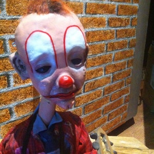 May include: A creepy clown doll with red and white makeup, wearing a red and black jacket, holding a wooden guitar. The doll has a long, thin neck and a sad expression.