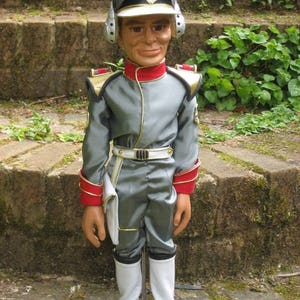 May include: A grey and white puppet dressed in a military uniform with red and gold trim. The puppet is wearing a white hat with a silver badge that says "WASP". The puppet is holding a white briefcase and has a microphone attached to its head.
