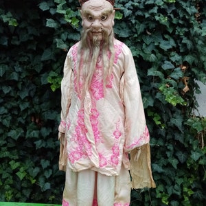 May include: A traditional Chinese puppet with a painted face and a long white beard. The puppet is wearing a pink and white robe with floral embroidery and a brown hat with Chinese characters.
