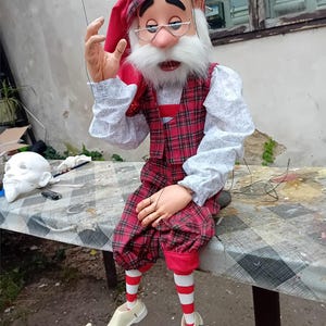 Könnte beinhalten: Eine rot-schwarze Karopuppen mit weißem Bart, roter Mütze und weiß-rot gestreiften Strümpfen. Die Puppe trägt ein weißes Hemd mit einer rot-schwarzen Karoweste. Die Puppe sitzt auf einem Tisch mit den Händen auf dem Schoß.