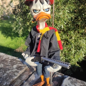 May include: A puppet of a duck wearing a red hat, black jacket, blue jeans, and a red and yellow scarf. The duck is holding a silver shotgun.