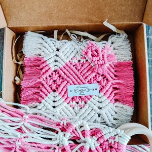 May include: A pink and white macrame wall hanging with a heart design and a small wooden ring. The hanging is packaged in a brown cardboard box with white tissue paper. The tag on the hanging reads "All handmade Design".