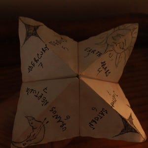 May include: A folded paper fortune teller featuring hand-drawn illustrations of celestial bodies including the sun, stars, and a crescent moon. The paper is inscribed with the names of planets such as Mercury, Venus, Earth, Mars, Saturn, Uranus, and Neptune.