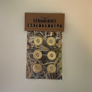Puede incluir: Un paquete de seis tapones de botella de oro en forma de casquillo de escopeta con un fondo de camuflaje. El paquete tiene una etiqueta marrón con el texto "LESIGOURNEY" y una estrella.