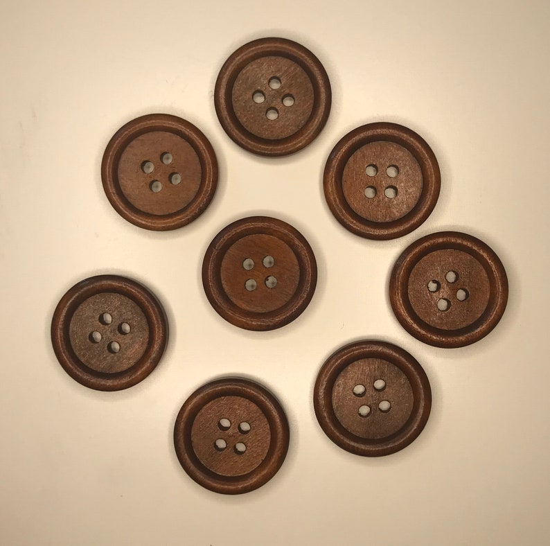 May include: A collection of eight round, dark brown wooden buttons with four holes each. The buttons are arranged in a circular pattern against a white background. These buttons are suitable for sewing and craft projects.