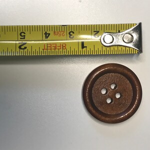 May include: A wooden button with four holes next to a yellow measuring tape. The button is brown and round. The measuring tape shows measurements in inches and feet.