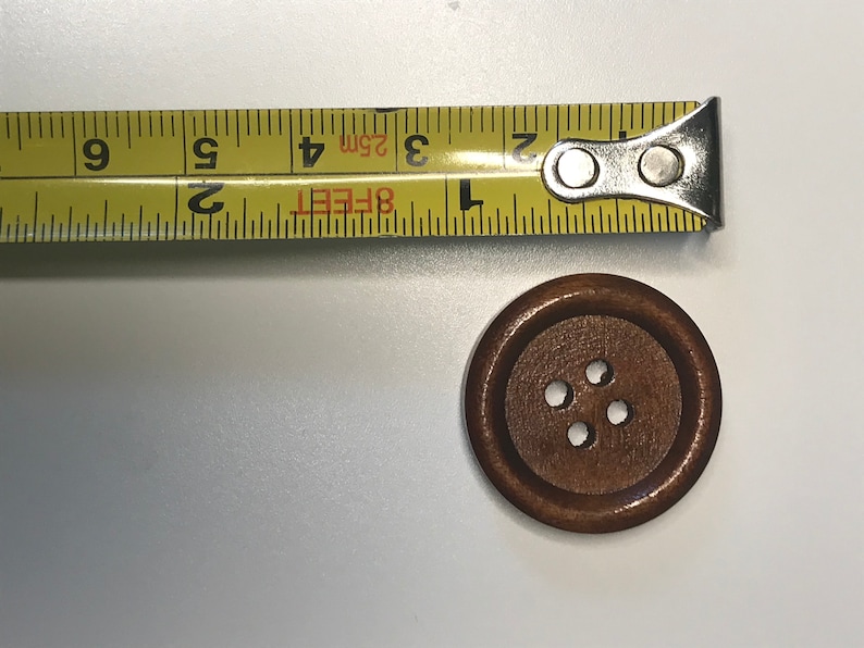 May include: A wooden button with four holes next to a yellow measuring tape. The button is brown and round. The measuring tape shows measurements in inches and feet.