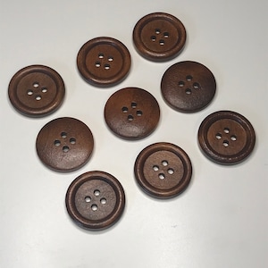 May include: A collection of nine dark brown, round wooden buttons with four holes each. The buttons are arranged on a white surface, showcasing their simple, classic design. These buttons are suitable for various sewing and craft projects.
