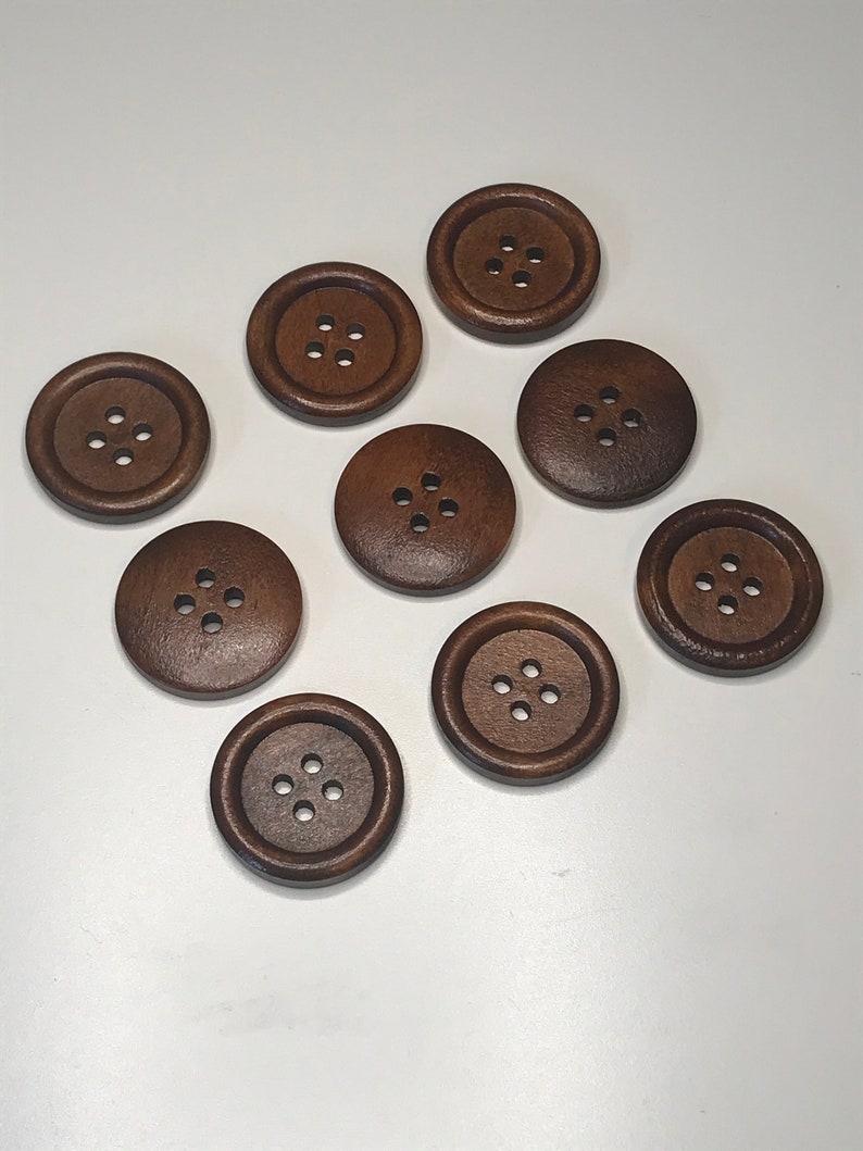 May include: A collection of nine dark brown, round wooden buttons with four holes each. The buttons are arranged on a white surface, showcasing their simple, classic design. These buttons are suitable for various sewing and craft projects.