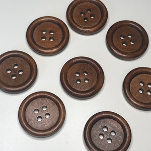 May include: A collection of eight large, round, dark brown wooden buttons. Each button has four small holes in the center for sewing. The buttons are evenly spaced against a white background, showcasing their rich color and simple design.