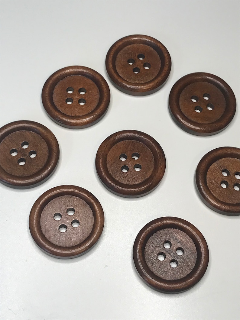 May include: A collection of eight large, round, dark brown wooden buttons. Each button has four small holes in the center for sewing. The buttons are evenly spaced against a white background, showcasing their rich color and simple design.
