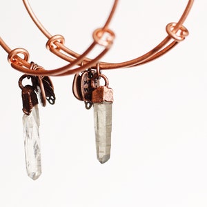 May include: Two copper wire bracelets with crystal charms. The charms are clear quartz crystals with copper accents. The bracelets are connected together by a copper wire.