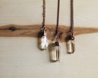 May include: Three clear quartz crystal pendants hanging from copper chains on a light wood background.