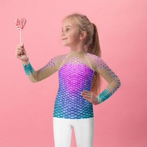 May include: A young person wearing a long-sleeved shirt with a mermaid scale pattern in shades of teal, blue, purple, and pink. The shirt is sheer and has a mesh overlay. The person is holding a red and white candy cane.