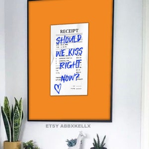 Puede incluir: Un recibo blanco con tinta azul que dice "SHOULD WE KISS RIGHT NOW?" con un coraz&oacute;n dibujado en la parte inferior. El recibo est&aacute; fijado a un fondo naranja brillante.