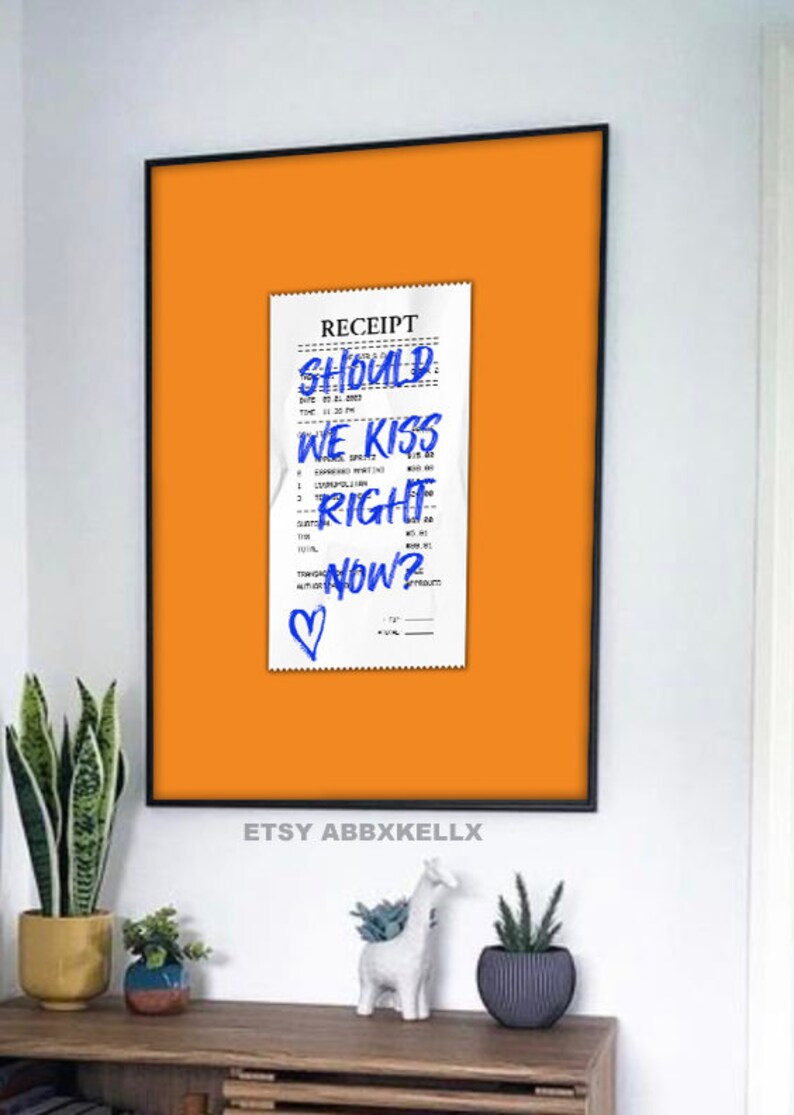 Puede incluir: Un recibo blanco con tinta azul que dice "SHOULD WE KISS RIGHT NOW?" con un coraz&oacute;n dibujado en la parte inferior. El recibo est&aacute; fijado a un fondo naranja brillante.