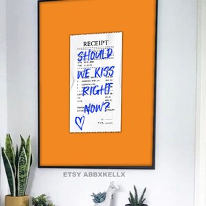 May include: A framed print of a receipt with the words "SHOULD WE KISS RIGHT NOW?" written in blue ink. A small heart is drawn at the bottom of the receipt. The background is a bright orange color.