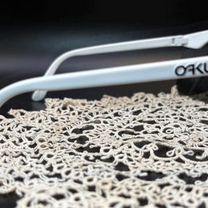 Może przedstawiać: Białe okulary przeciwsłoneczne Oakley z czarnym napisem na zauszniku. Okulary przeciwsłoneczne spoczywają na kremowej, ażurowej tkaninie na ciemnym tle. Ostrość skupia się na oprawkach okular&oacute;w i nazwie marki.