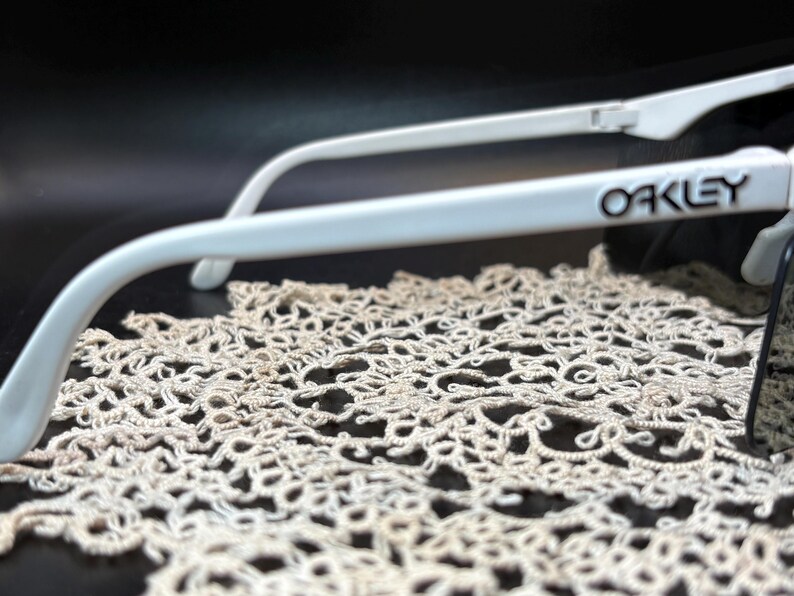 Może przedstawiać: Białe okulary przeciwsłoneczne Oakley z czarnym napisem na zauszniku. Okulary przeciwsłoneczne spoczywają na kremowej, ażurowej tkaninie na ciemnym tle. Ostrość skupia się na oprawkach okular&oacute;w i nazwie marki.